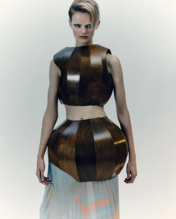 A model poses in a futuristic, geometrically shaped wooden crop top and skirt with a pleated pastel skirt in a studio setting.