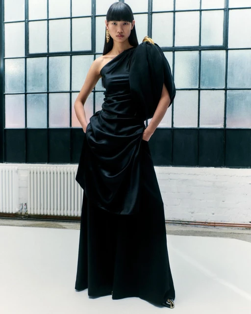 Elegant Asian woman poses in a black one-shoulder satin gown with gold earrings in a bright studio with a large window.