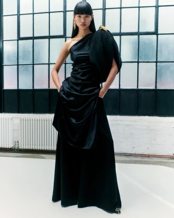 Elegant Asian woman poses in a black one-shoulder satin gown with gold earrings in a bright studio with a large window.