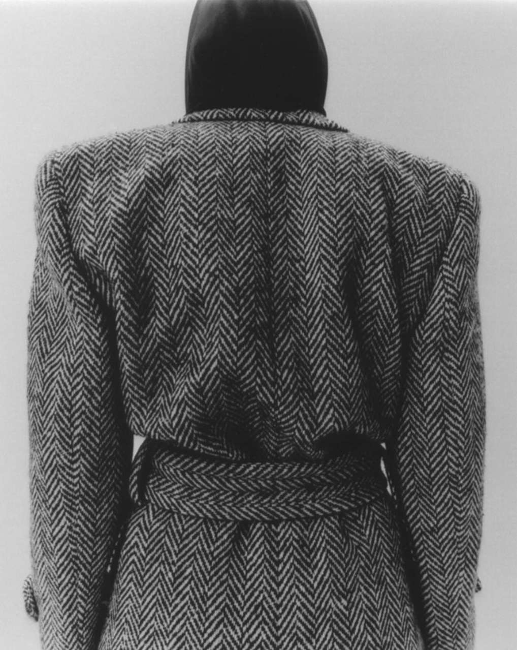 Back view of a person wearing a herringbone tweed coat with a belt, standing against a neutral background in a studio setting.