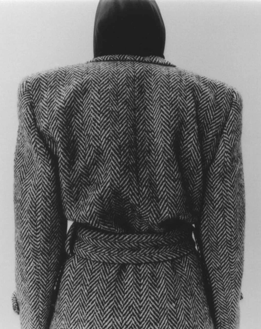 Back view of a person wearing a herringbone tweed coat with a belt, standing against a neutral background in a studio setting.