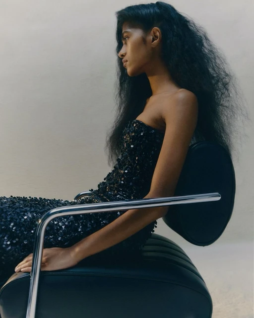 A dark-skinned woman with long curly dark hair sits in a black leather chair wearing a strapless black sequined dress in a studio setting.