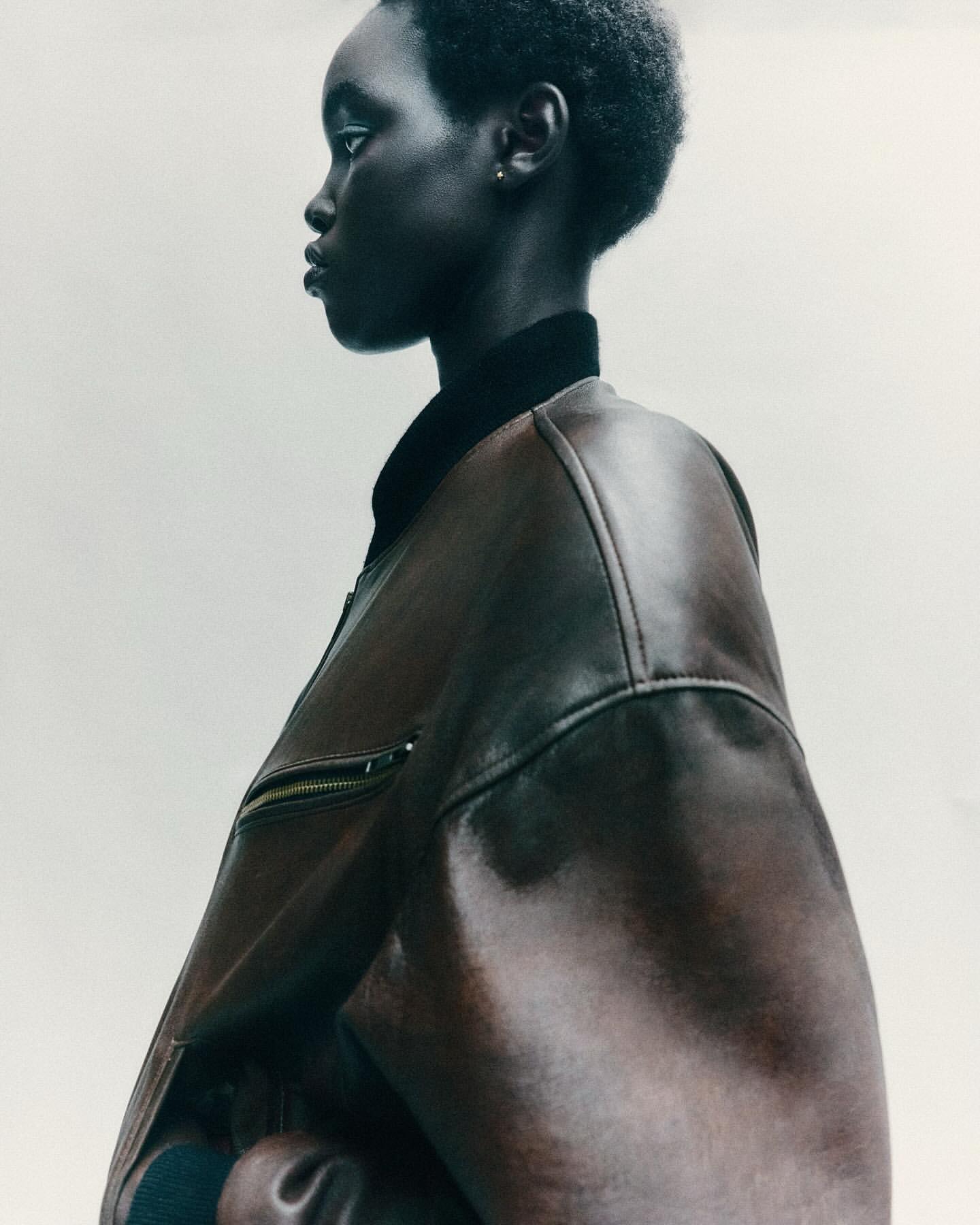 Portrait of Black woman in leather jacket, side view against white backdrop, showcasing fashion and beauty in a minimalist setting.