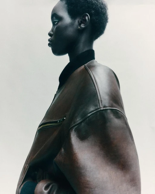 Portrait of Black woman in leather jacket, side view against white backdrop, showcasing fashion and beauty in a minimalist setting.