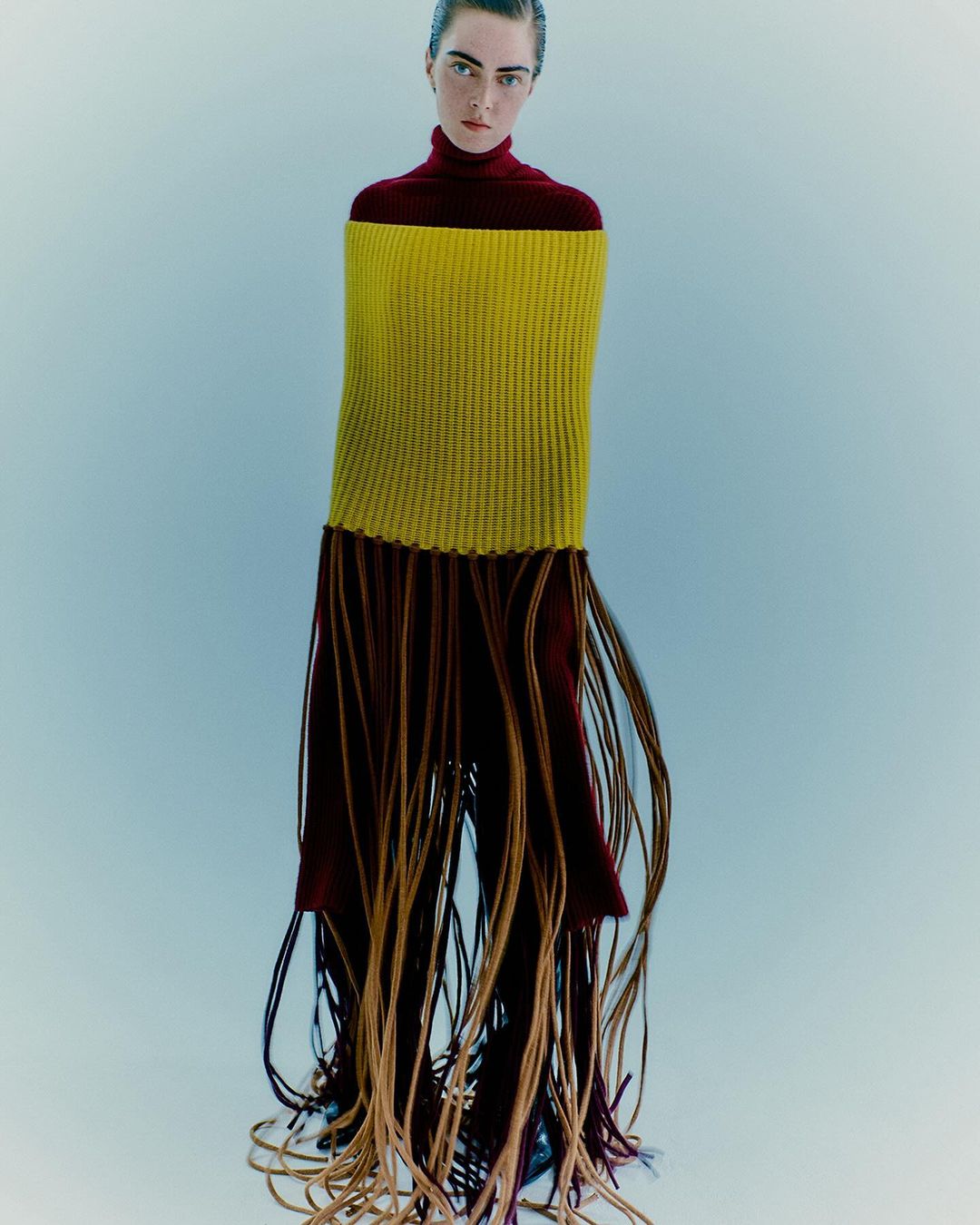 Fashion shot featuring an adult female in a red turtleneck and a yellow textile piece with fringed detailing, posing in a studio setting.