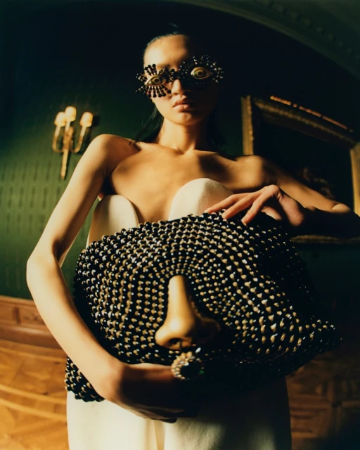 Woman poses indoors with beaded bag with nose detail and beaded eye-glasses. Dark, moody lighting and surreal fashion elements.