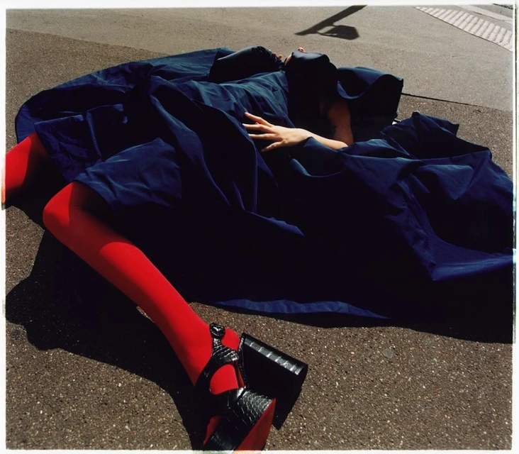 Woman lying on asphalt in a dark blue fabric dress, red tights, and black platform heels; overhead shadow. Fashion editorial.