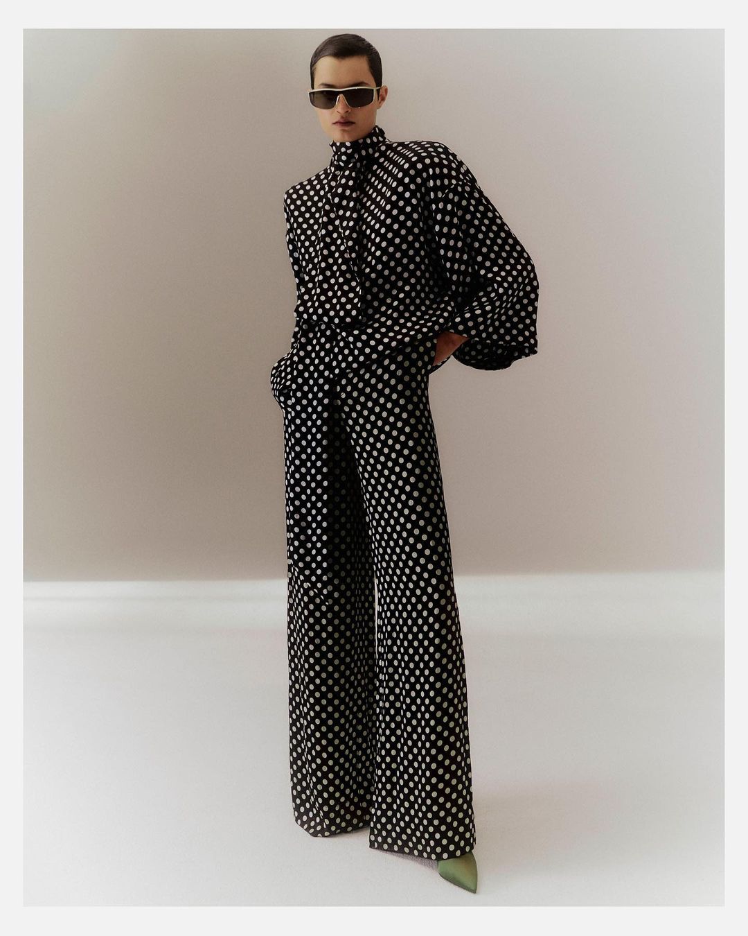 A woman in a black and white polka dot pantsuit stands in a photo studio wearing dark glasses and green heeled shoes.