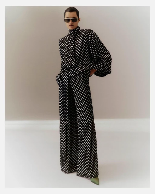 A woman in a black and white polka dot pantsuit stands in a photo studio wearing dark glasses and green heeled shoes.
