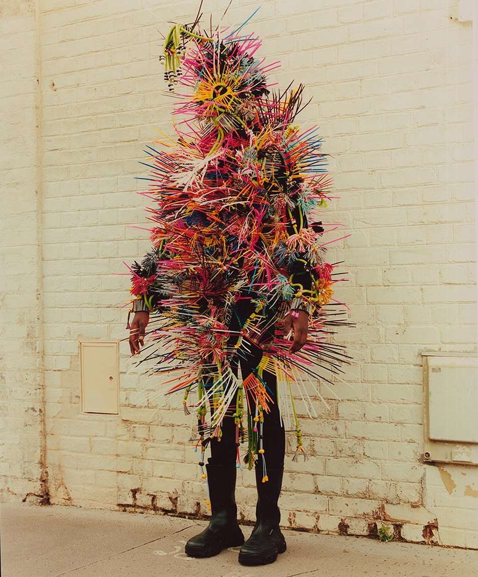 Person in a colorful, spiky costume stands against a white brick wall, creating a vibrant and eye-catching visual statement.