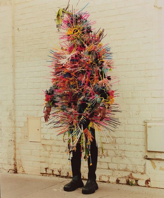 Person in a colorful, spiky costume stands against a white brick wall, creating a vibrant and eye-catching visual statement.