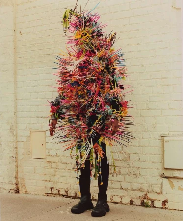 Person in a colorful, spiky costume stands against a white brick wall, creating a vibrant and eye-catching visual statement.