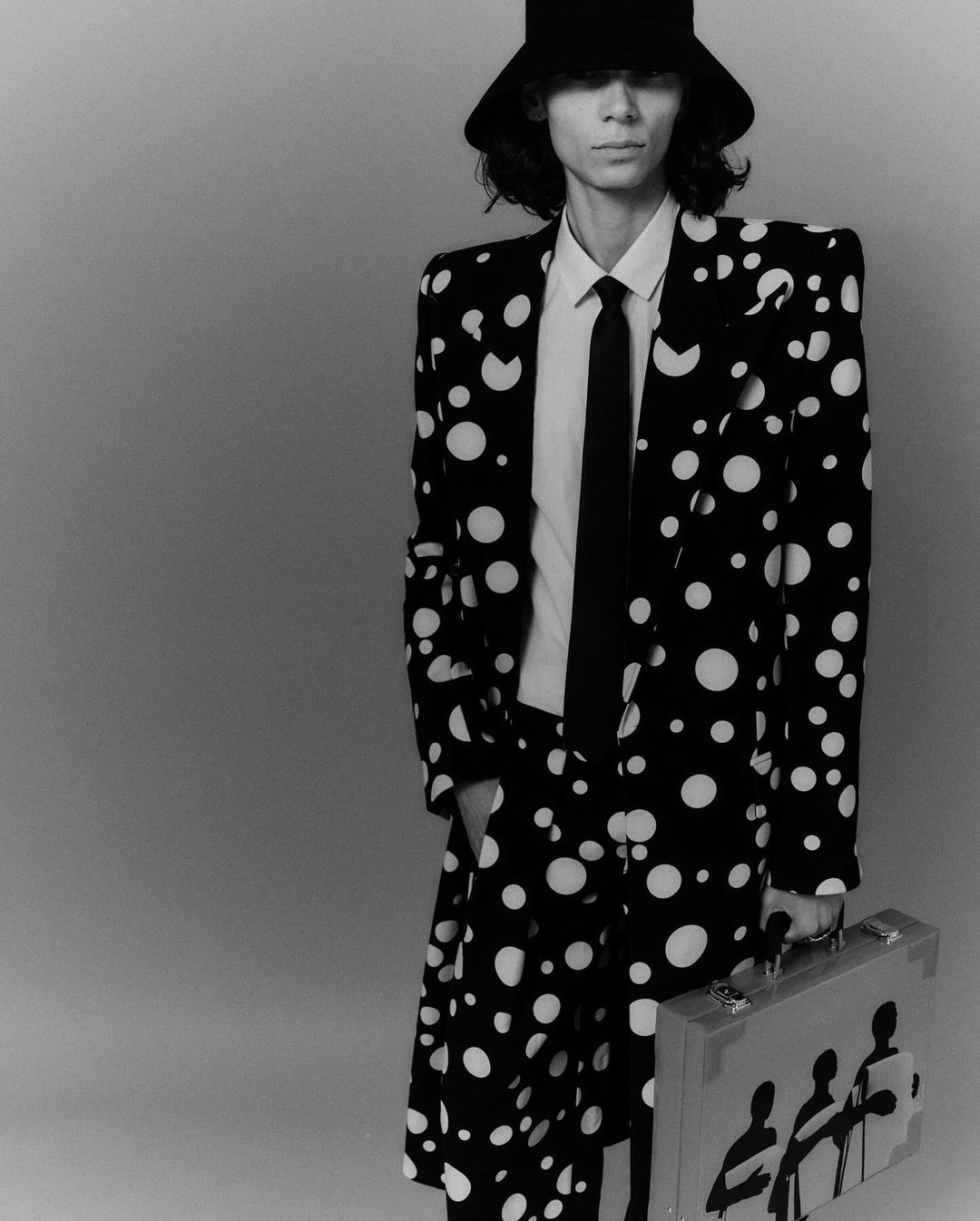A person wearing a polka dot suit and bucket hat stands in a studio, holding a briefcase with an illustration of silhouetted figures.
