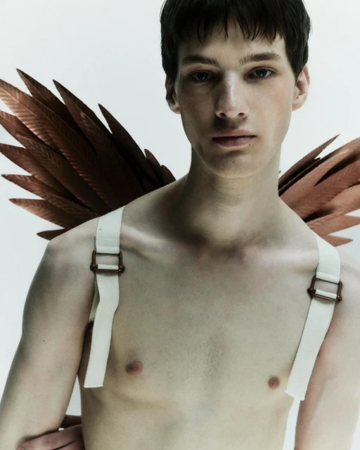 Striking portrait of a slender adult male with dark hair and light skin, wearing brown wings and white straps against a white background.