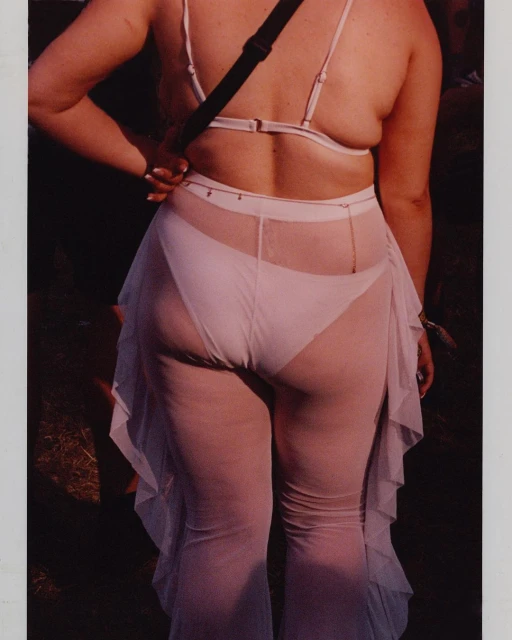 Back view of an adult female wearing a white bikini top and sheer white ruffled pants outdoors, visible against a dark background.
