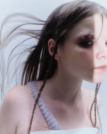 Close-up portrait of a woman with blowing hair, dark eye makeup, and a textured white strap top against a light background.
