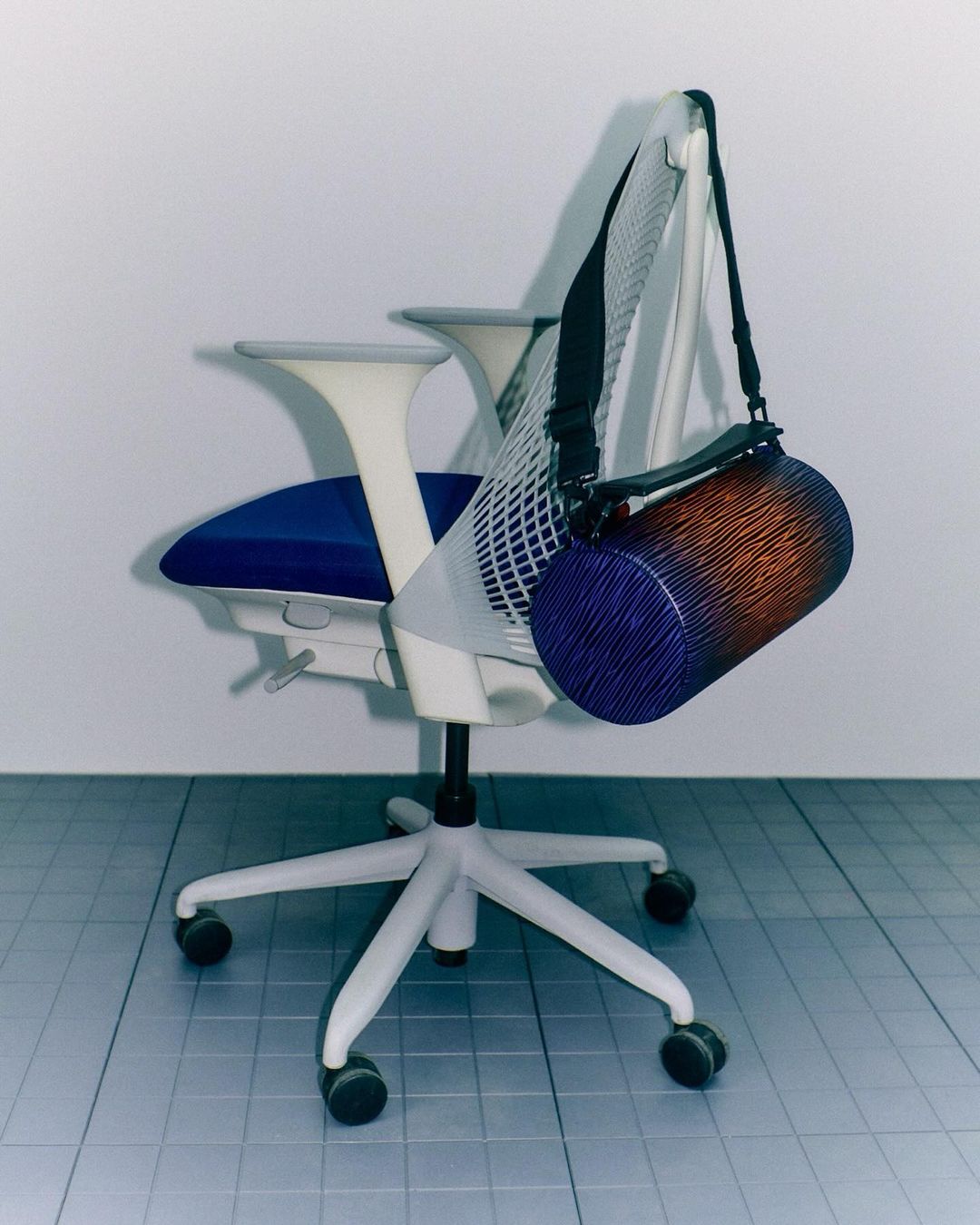Office chair with blue seat and gradient cylinder bag hanging, set in a simple studio with cool lighting and tiled floor.