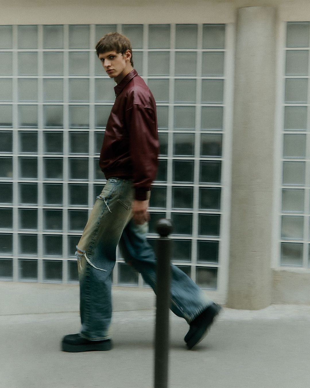 Male fashion model in burgundy bomber jacket and ripped jeans walking on city sidewalk with glass block wall in the background.