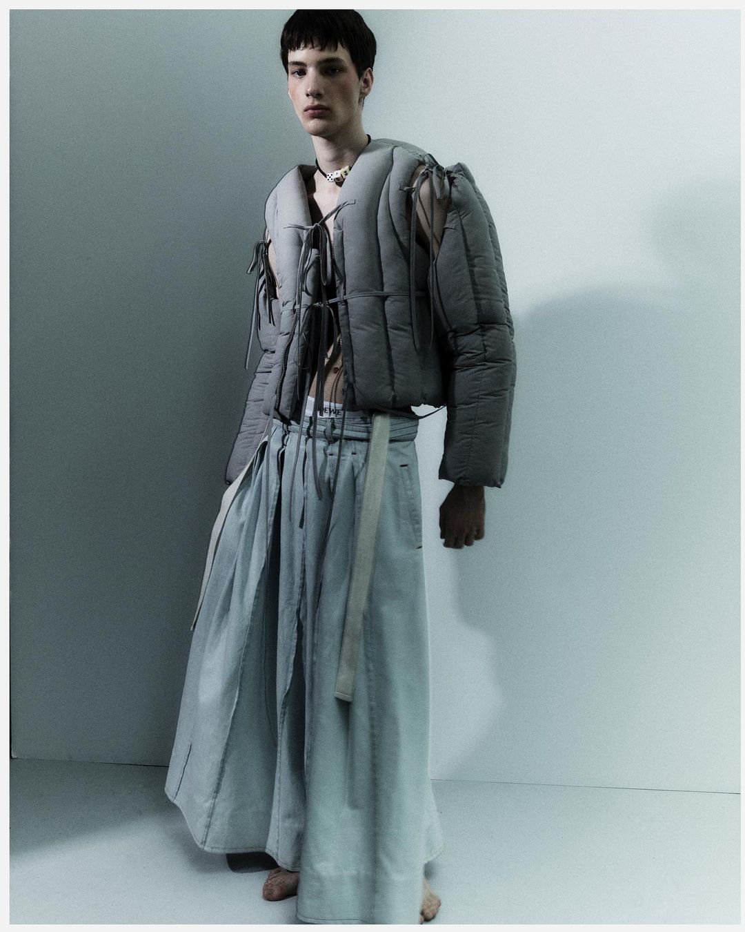 Fashionable young adult stands in a studio, wearing a gray puffer jacket and flowing skirt, complemented by subtle necklace detail.