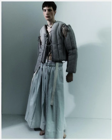 Fashionable young adult stands in a studio, wearing a gray puffer jacket and flowing skirt, complemented by subtle necklace detail.