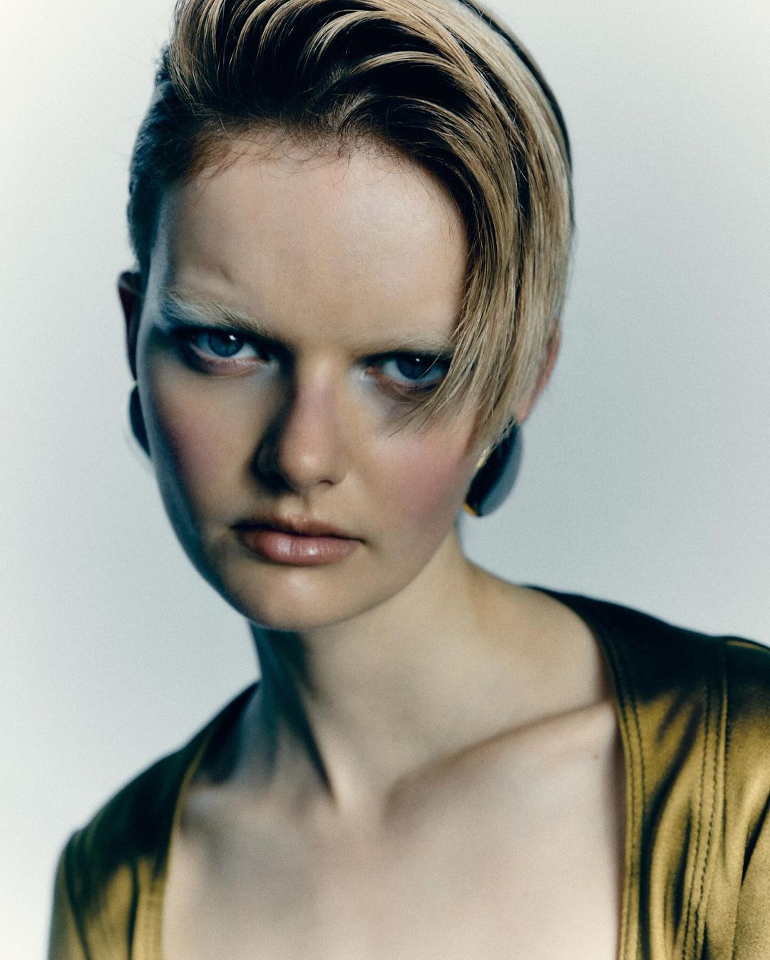 Close-up portrait of an adult female with blonde hair and blue eyes wearing a gold top against a light background, showcasing a beauty aesthetic.