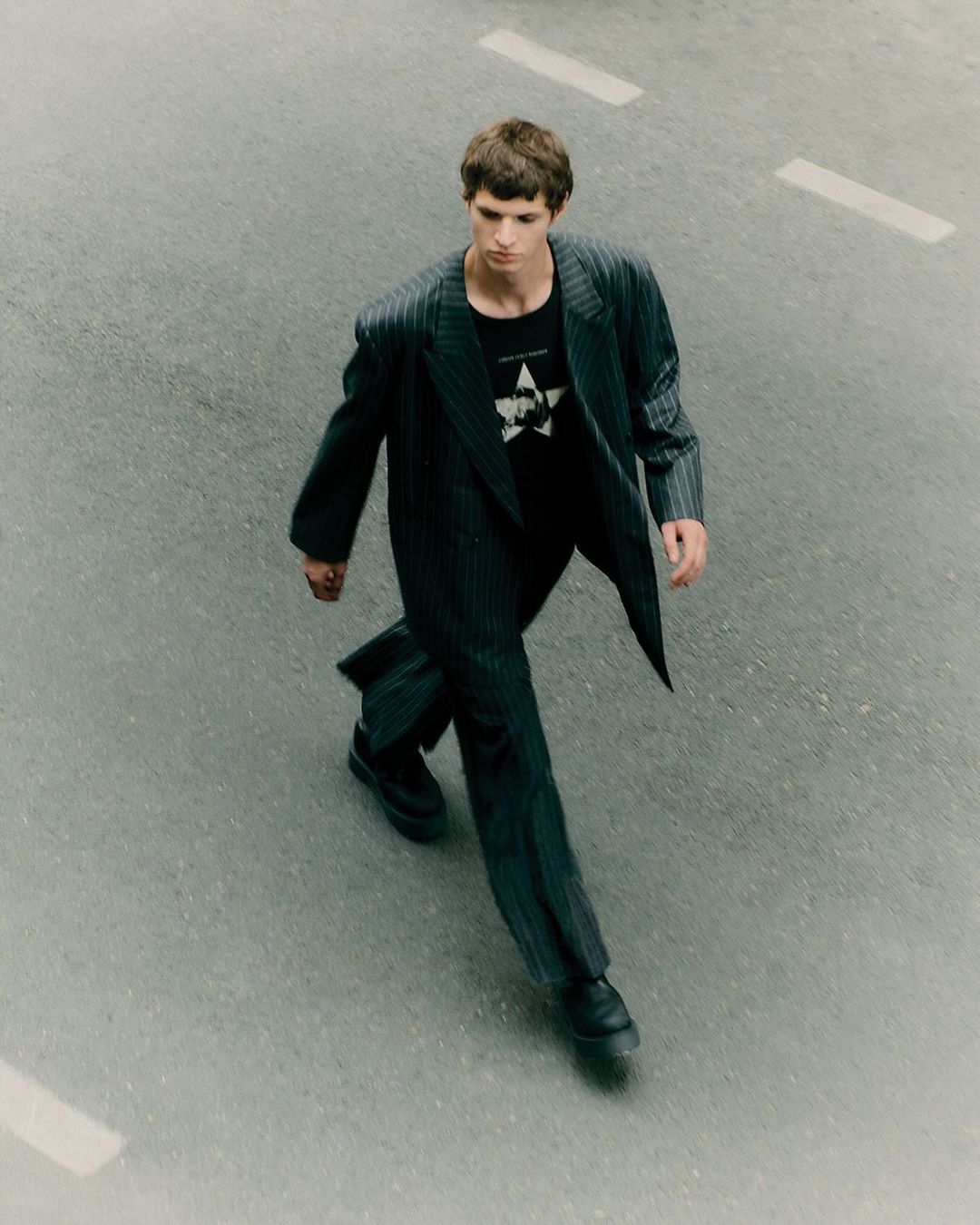 Young man walks on asphalt road wearing pinstripe suit and black t-shirt with star graphic, captured from an overhead perspective.