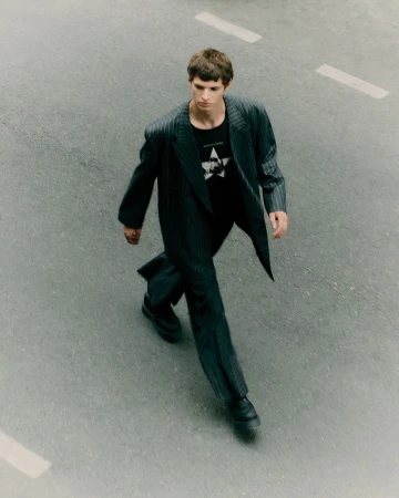 Young man walks on asphalt road wearing pinstripe suit and black t-shirt with star graphic, captured from an overhead perspective.