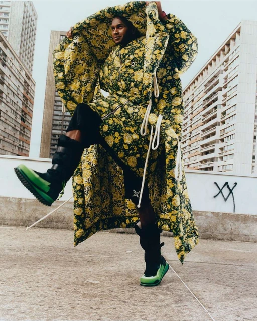 Editorial fashion shot of a person in a floral coat and green boots posing on a rooftop with high-rise buildings in the background.