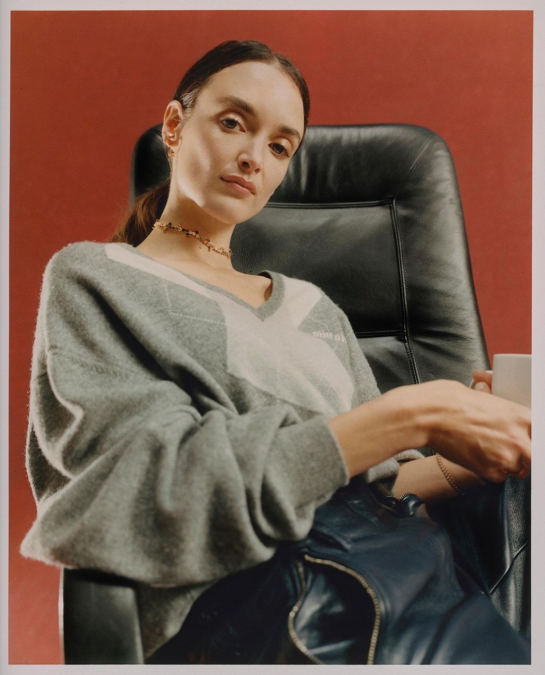 A young adult woman sits in a black chair holding a cup, wearing a gray sweater with white details against a solid red background.