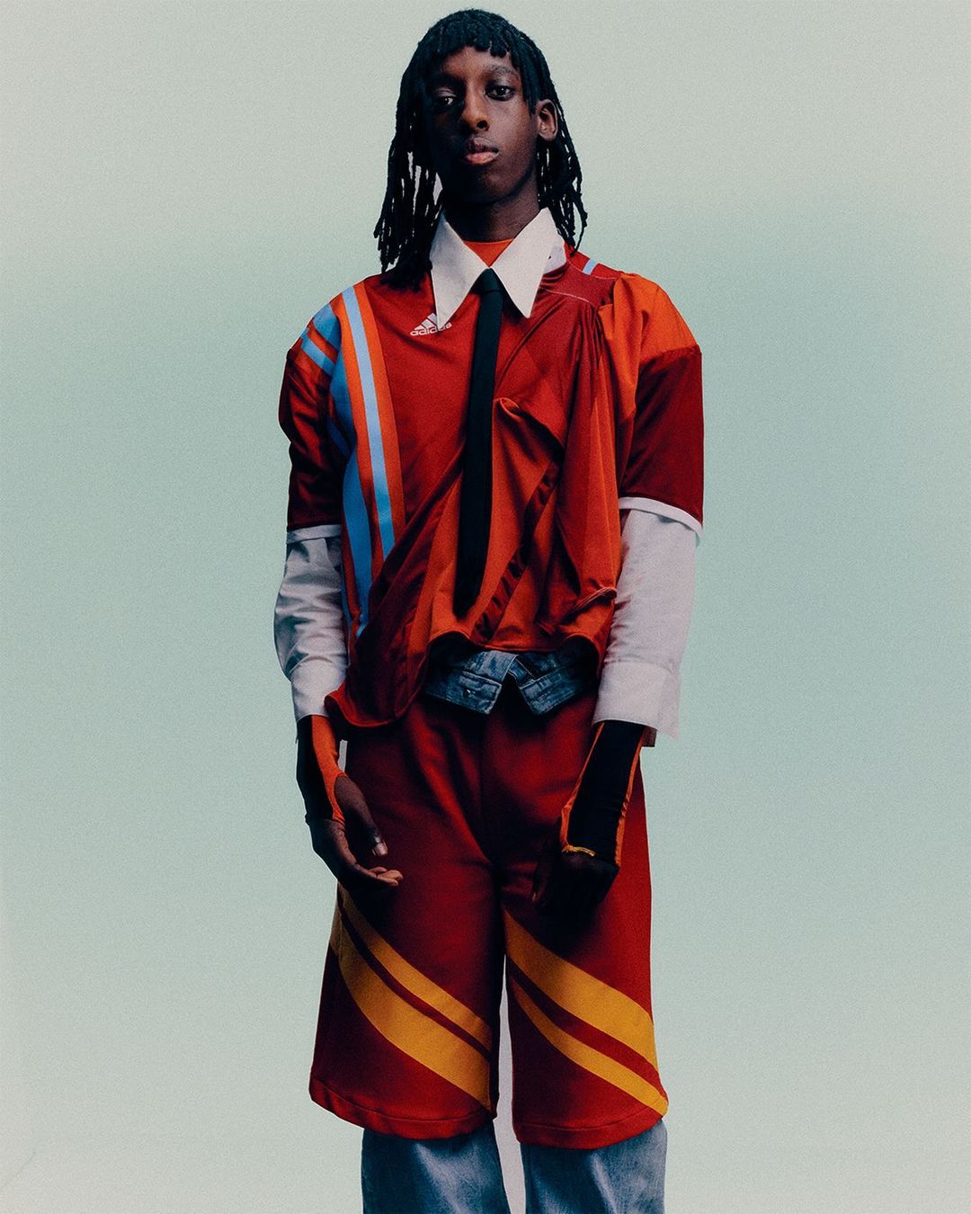 Fashionable young adult with dreadlocks posing in an orange, blue, and white athletic-inspired top, shorts, and jeans against a light blue background.