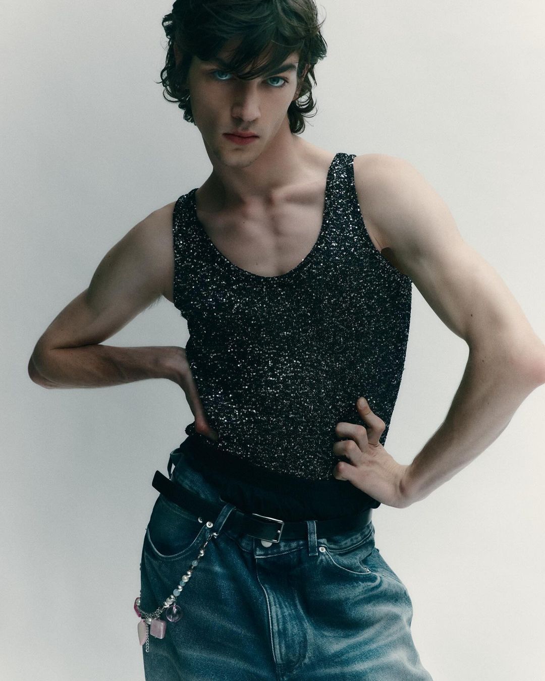 Young man in a sparkly tank top poses indoors with hands on his hips, showcasing a contemporary fashion style against a neutral backdrop.