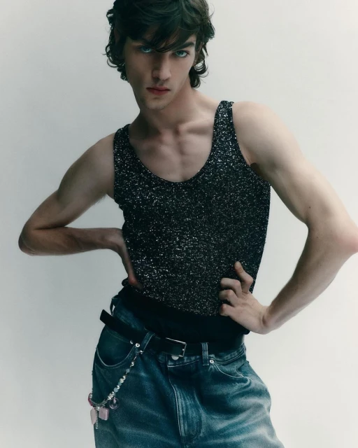 Young man in a sparkly tank top poses indoors with hands on his hips, showcasing a contemporary fashion style against a neutral backdrop.