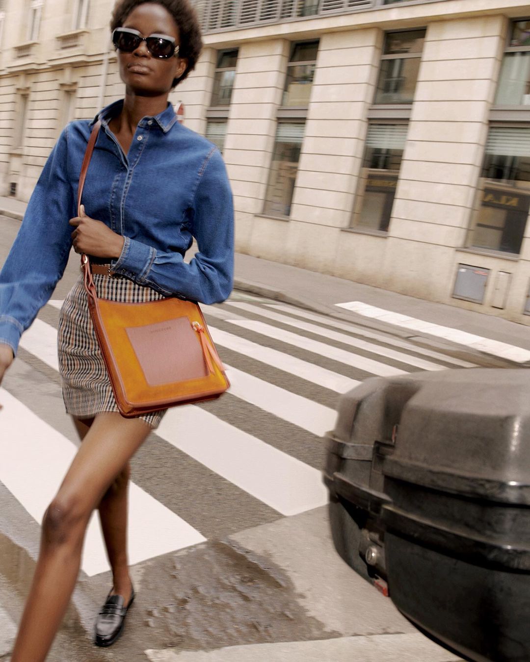 Woman in denim shirt and plaid skirt crossing the street carrying a brown bag with city buildings in the background.