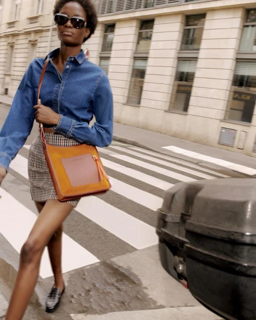 Woman in denim shirt and plaid skirt crossing the street carrying a brown bag with city buildings in the background.