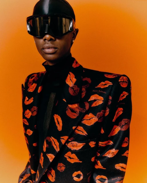 Portrait of an adult woman wearing oversized sunglasses and a blazer patterned with lipstick marks against a solid orange backdrop.