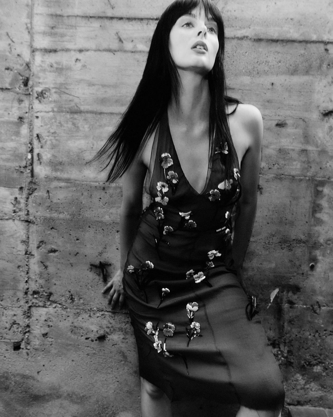 Monochrome portrait of a stylish woman with long dark hair wearing a floral dress, posing against a textured concrete wall.