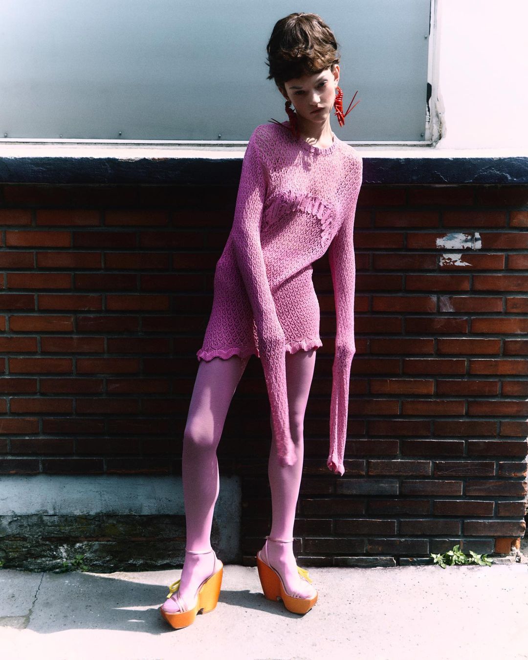 Fashionable young adult woman in a pink knit dress and pink tights poses against a brick wall wearing platform shoes.