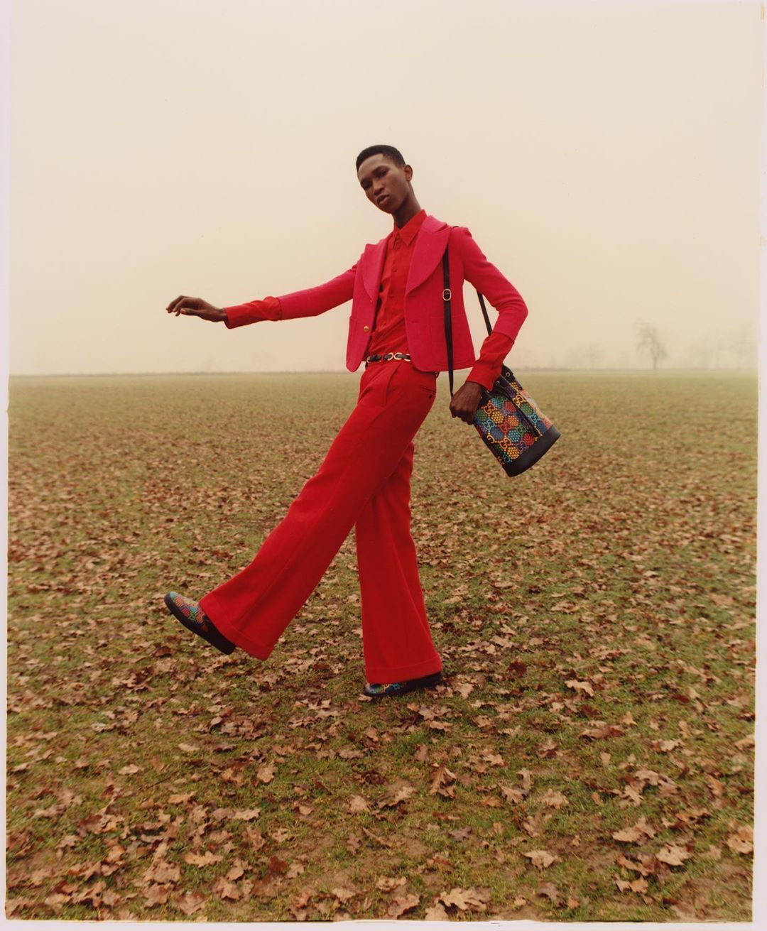 A stylish adult in a red jacket and pants strides across a grassy field with fallen leaves, carrying a patterned tote bag, captured in a medium shot.