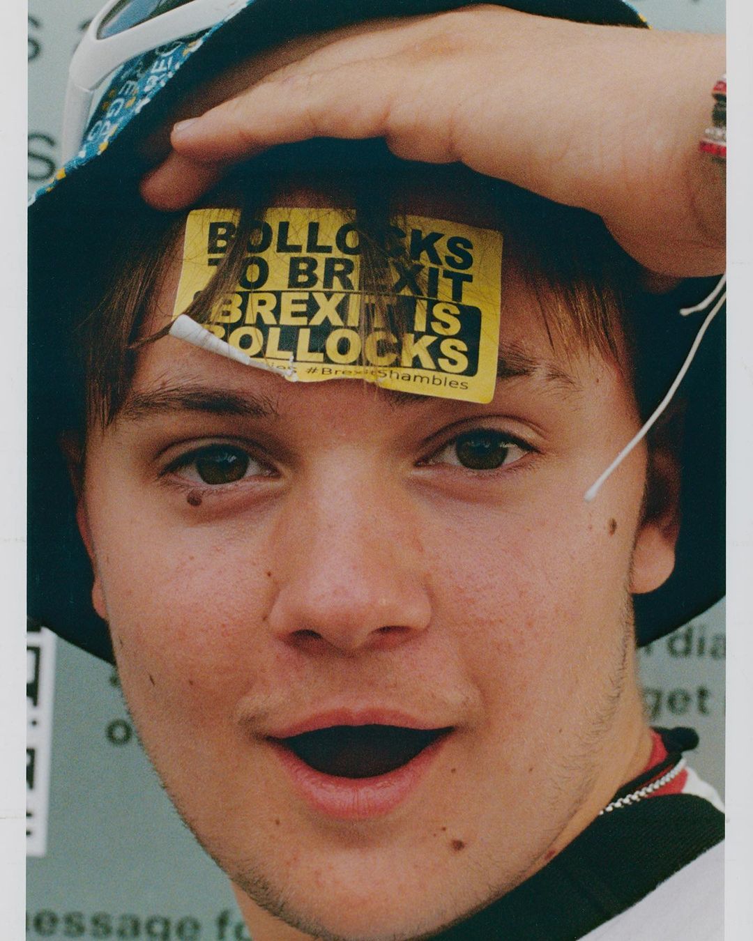 Close-up portrait of a young adult with a political sticker on their forehead, wearing a patterned bucket hat and a striped shirt.