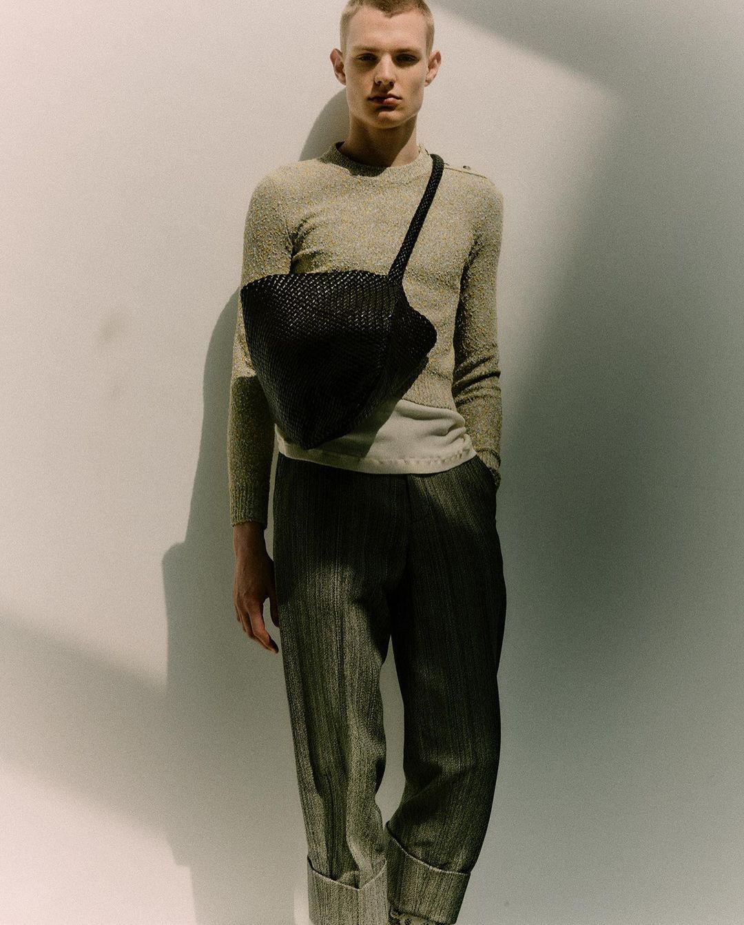 Blonde male model in a neutral sweater and pleated pants poses against a white wall with a black woven shoulder bag.