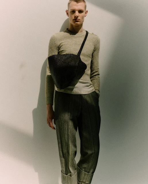 Blonde male model in a neutral sweater and pleated pants poses against a white wall with a black woven shoulder bag.