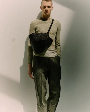 Blonde male model in a neutral sweater and pleated pants poses against a white wall with a black woven shoulder bag.