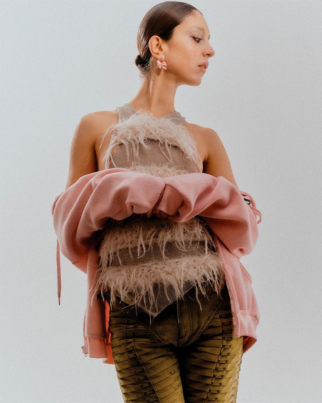 A woman poses in a beige top with feathers, green pants, and a pink jacket against a bright, neutral background in a fashion studio shoot.