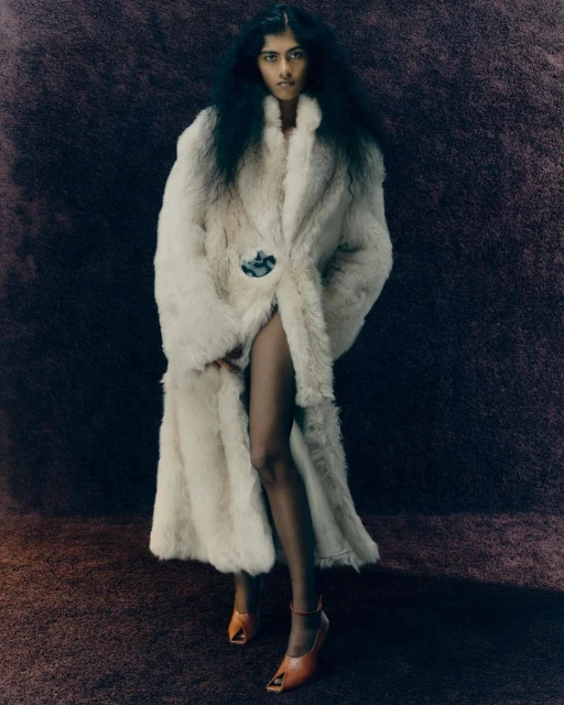 A woman with dark hair poses in a long, light-colored fur coat and tan shoes in a studio with a dark textured background.