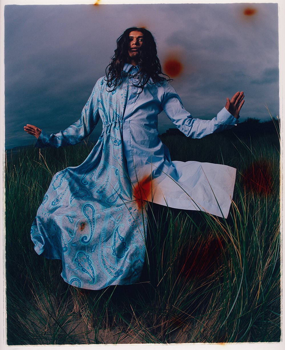 A single woman stands in a windswept field of tall grass wearing a long blue paisley print dress against a cloudy sky backdrop.