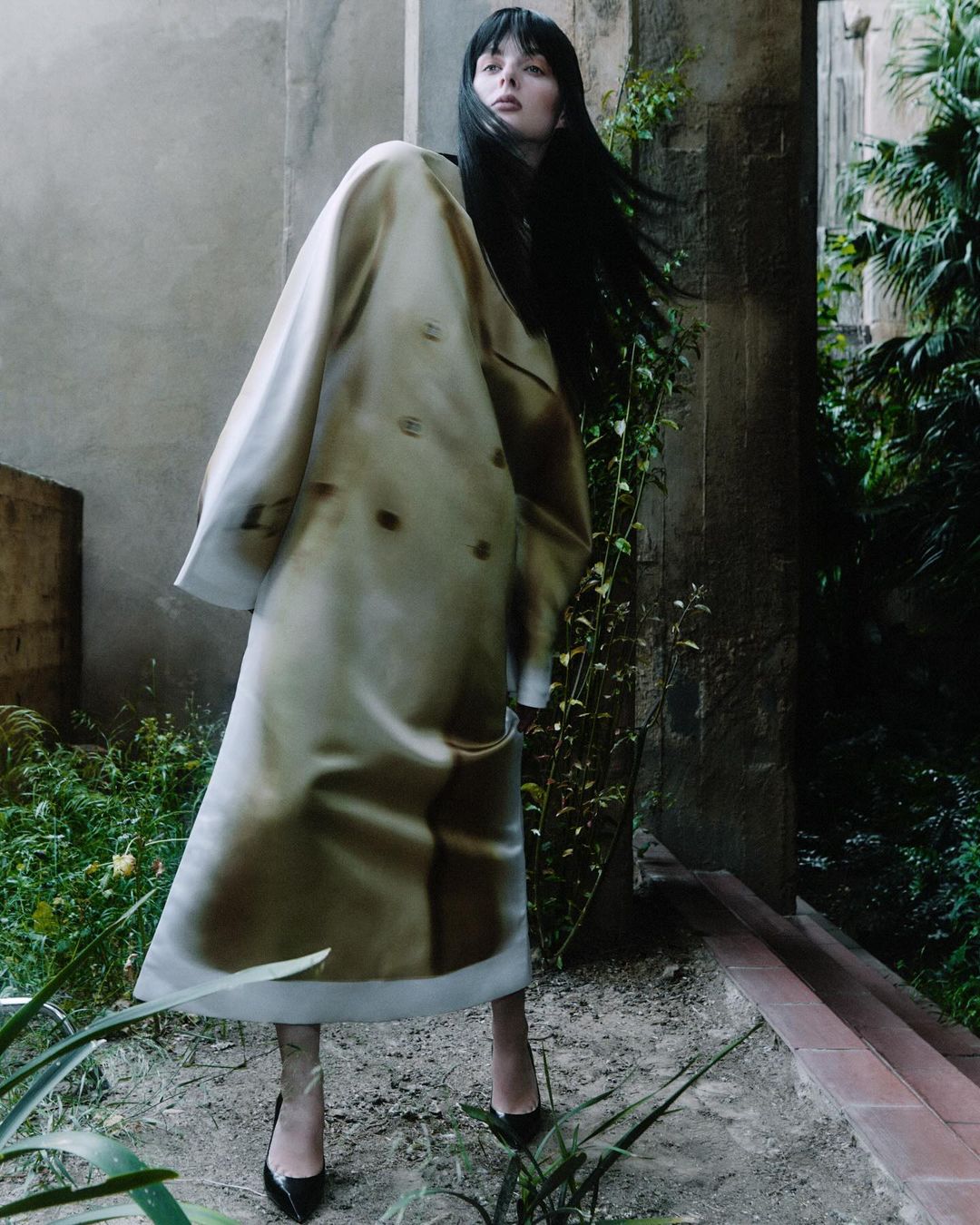 Model with long dark hair wearing a patterned coat and black heels standing outdoors near stone architecture