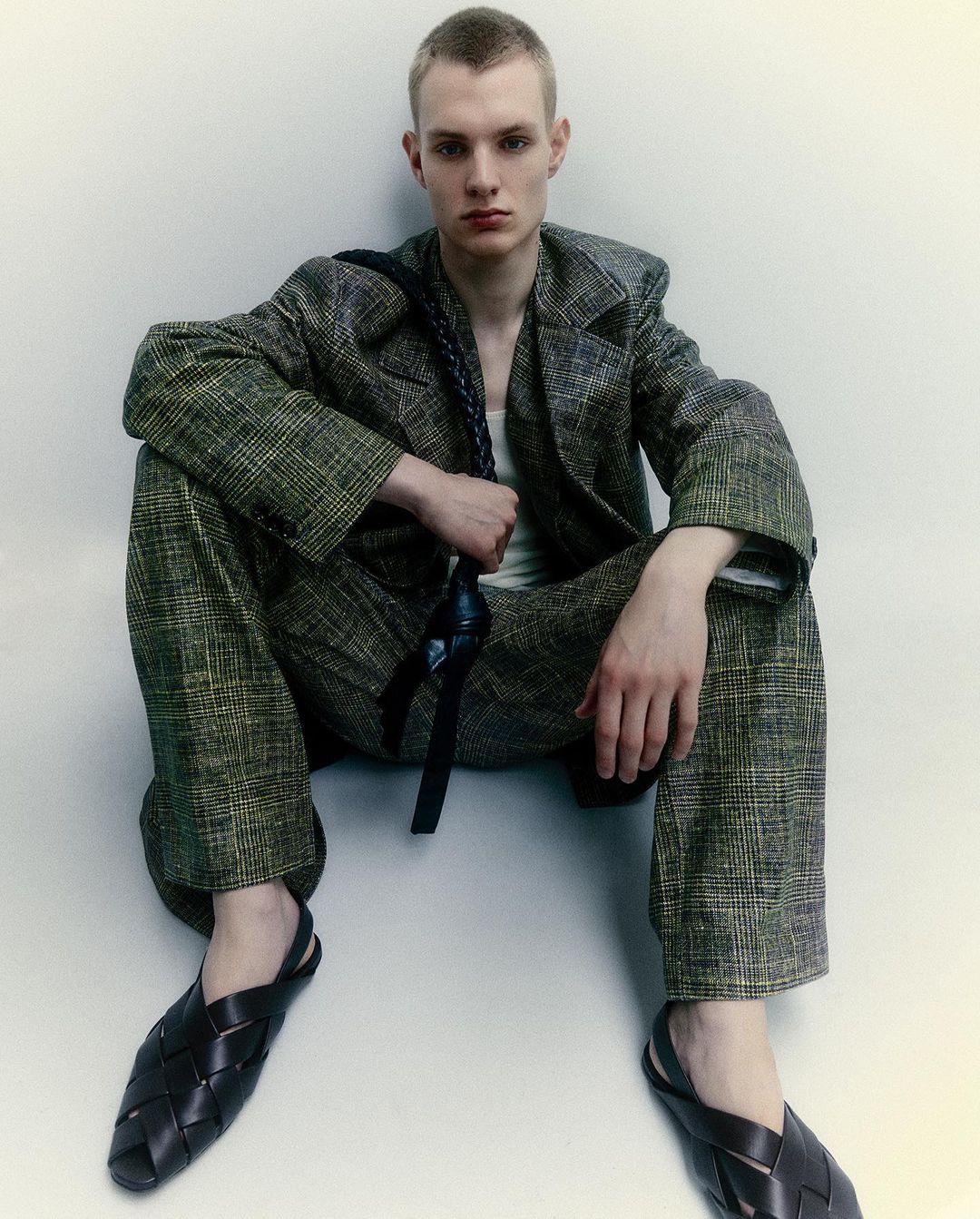 A fair-skinned young adult male model sits in a studio wearing a plaid suit, white tank top, and woven black leather slip-on shoes.