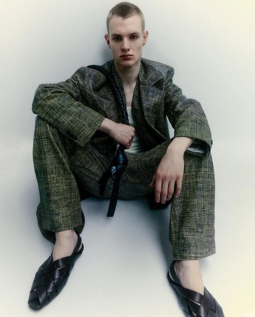 A fair-skinned young adult male model sits in a studio wearing a plaid suit, white tank top, and woven black leather slip-on shoes.