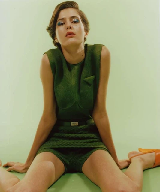Woman in green textured sleeveless dress and orange shoes sits on green background looking up at camera in a fashion studio shot.