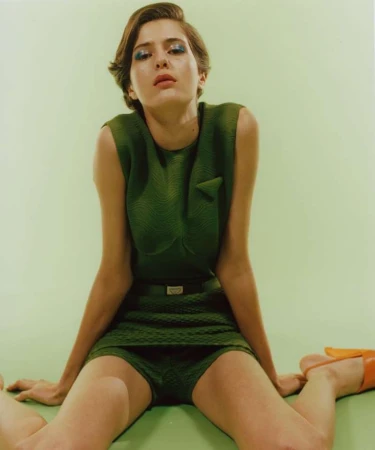 Woman in green textured sleeveless dress and orange shoes sits on green background looking up at camera in a fashion studio shot.
